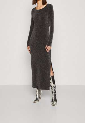 Kobieta ubrana w czarną, długo rękawową, błyszczącą sukienkę midi z bocznym rozcięciem, w połączeniu z błyszczącymi srebrnymi kozakami za kolano na jednolitym tle.