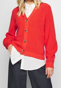 Cárdigan de punto rojo con escote en V, seis botones marrones, puños acanalados y un diseño texturizado. Llevado sobre una camisa blanca con cuello.