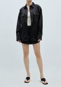 Schwarze satinierte Übergrößenshirt mit zwei Fronttaschen, kombiniert mit einem eng anliegenden schwarzen Minirock und schwarzen Absatzsandalen. Glatte Textur, minimalistisches Design.