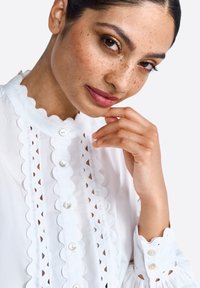 Chemisier blanc avec un décolleté festonné et un devant à boutons, comportant des découpages brodés. Manches longues avec des poignets froncés et un détail de bouton.