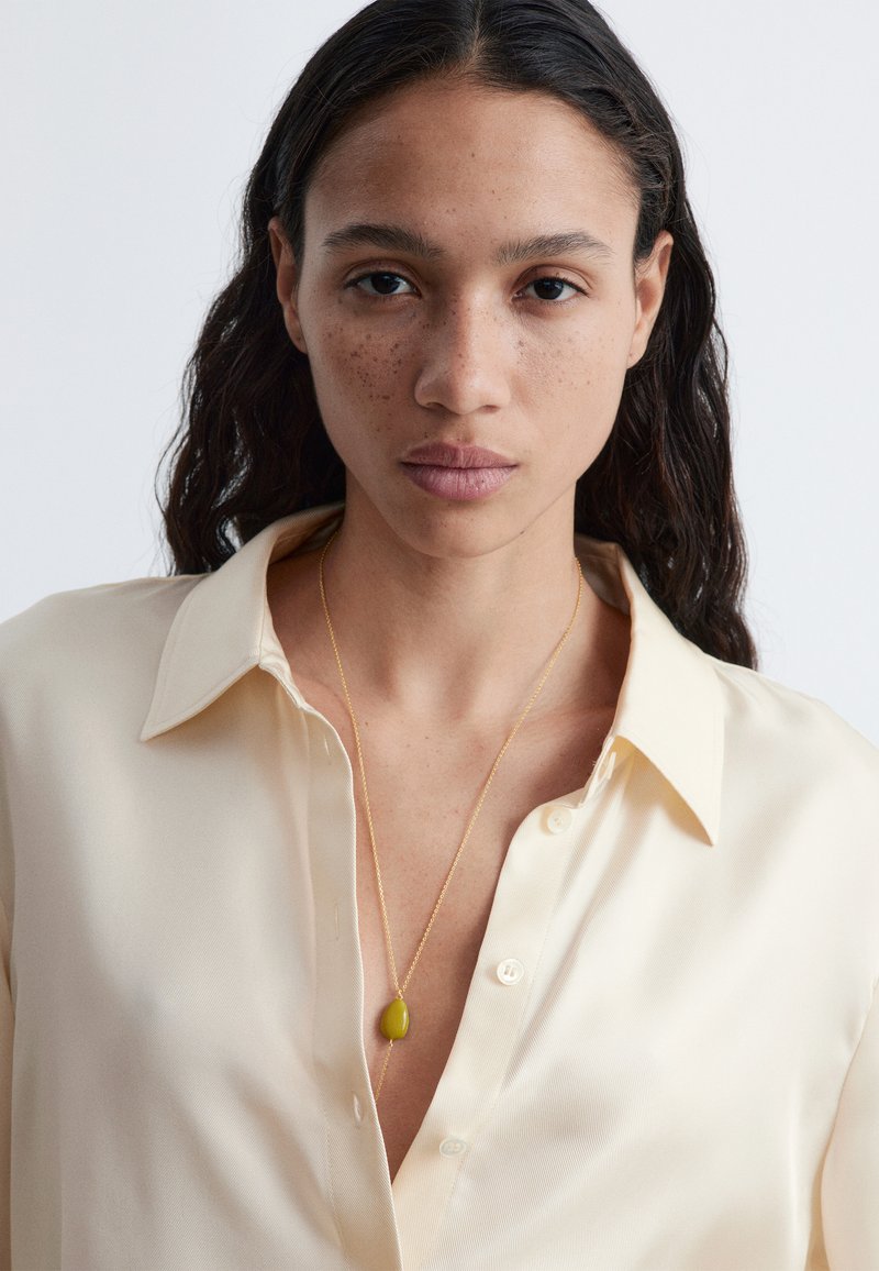 Jeune femme aux longs cheveux bruns portant un chemisier en soie crème et un collier en or avec un pendentif en pierre jaune sur un fond uni.