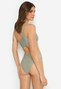 Σετ μπικίνι σε πράσινο χρώμα με τοπ ενός ώμου που διαθέτει άνοιγμα και σλιπ ψηλόμεσο. Ομαλή υφή και μινιμαλιστικός σχεδιασμός.