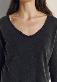 Ženski zgornji del trupa, oblečen v temno sivo majico z dolgimi rokavi in izpostavljenim V-izrezom z neobdelanimi robovi, na katerem so vidni ključnički in razpršene kožne pegice.