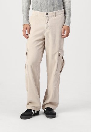 Beiga kordflor cargobyxor med två sidfickor, rak ben, subtil ribbad textur, kombinerat med svarta sneakers med vita accent.
