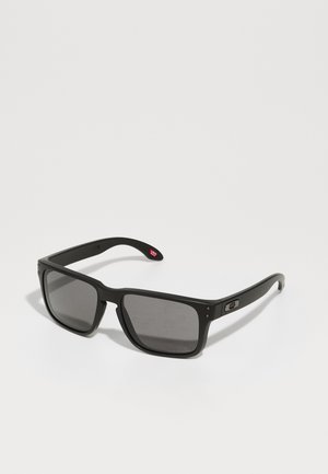 Occhiali da sole rettangolari neri con lenti scure, montatura opaca e dettagli del logo discreti sul lato. Presentano aste resistenti e un design leggero.