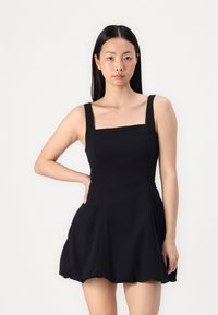 Svart ermeløs kjole med firkantet hals, tettsittende bodice, flagrende skjørt og minimale sømmer. Laget av glatt stoff med myk tekstur.
