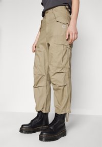 Beige cargobyxor med flera fickor, elastiska muddar och en avslappnad passform, parat med svarta snörstövlar med tjock sula.