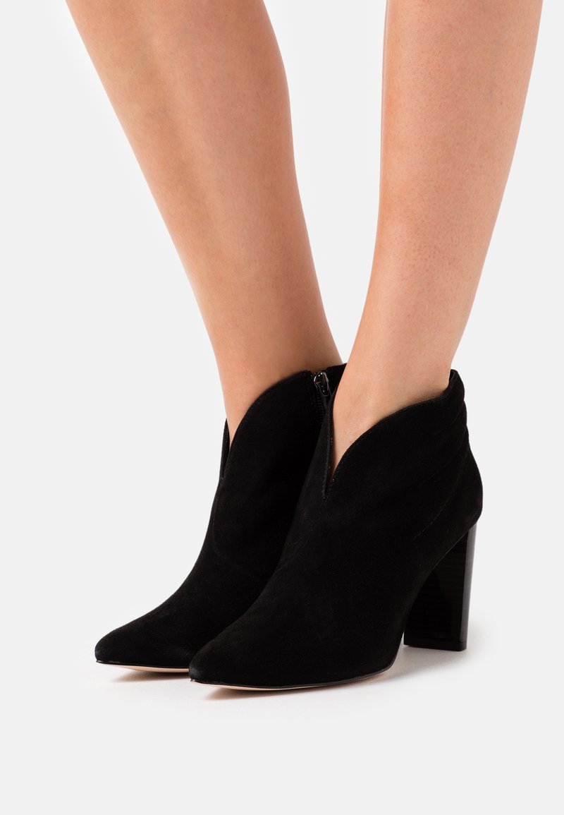 Bottes chevilles en daim noir avec bouts pointus, talons blocs hauts et ouvertures supérieures en forme de V, portées sur des jambes nues.