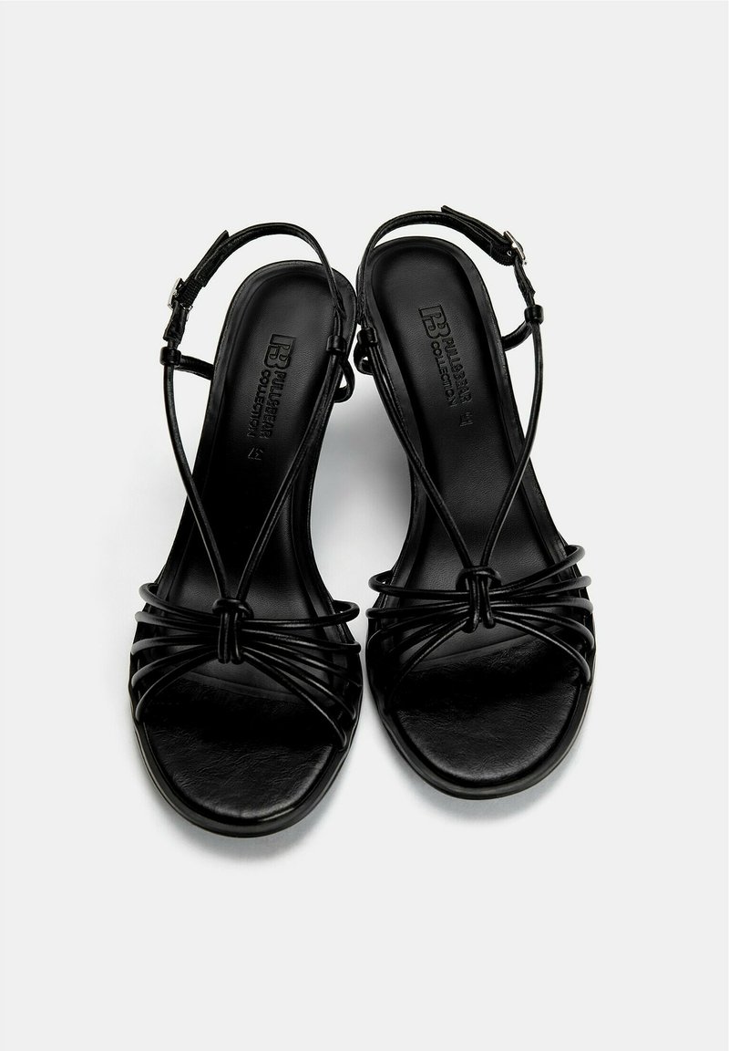 Sandalias negras con un acabado suave, que cuentan con intrincadas correas entrelazadas y una correa ajustable en el tobillo, sobre una suela plana.