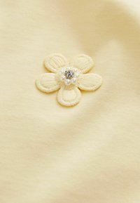 Gelber Stoff mit einer Blumenapplikation, die gestickte Blütenblätter, eine zentrale dekorative Perle und perlenartige Akzente aufweist. Glatte Textur.