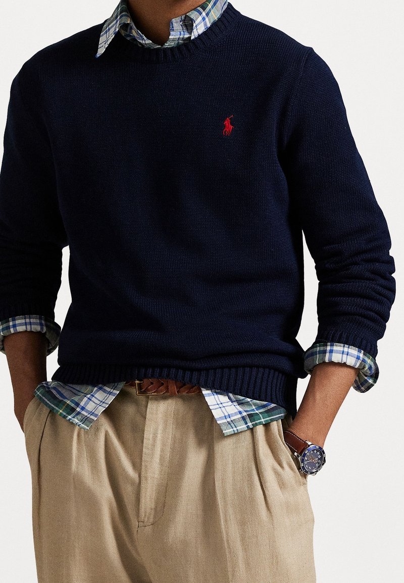 Homme portant un pull bleu marine par-dessus une chemise à carreaux bleue et verte à boutons, pantalon beige, ceinture tressée marron et montre-bracelet.