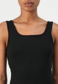 Zwarte geribbelde tanktop met een vierkante halslijn en dunne bandjes, met een textuurstof en een golvende rand langs de halslijn.