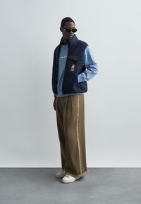 Ναυτικό γιλέκο πάνω από μπλε ανοιχτό μακρυμάνικο πουκάμισο, συνδυασμένο με φαρδιά καφέ παντελόνια. Λευκά αθλητικά παπούτσια και γυαλιά ηλίου ολοκληρώνουν την εμφάνιση.