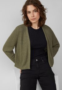 Gilet en tricot vert olive avec une ouverture devant et une texture côtelée, porté sur un haut noir avec de petites embellissements, associé à un jean noir.