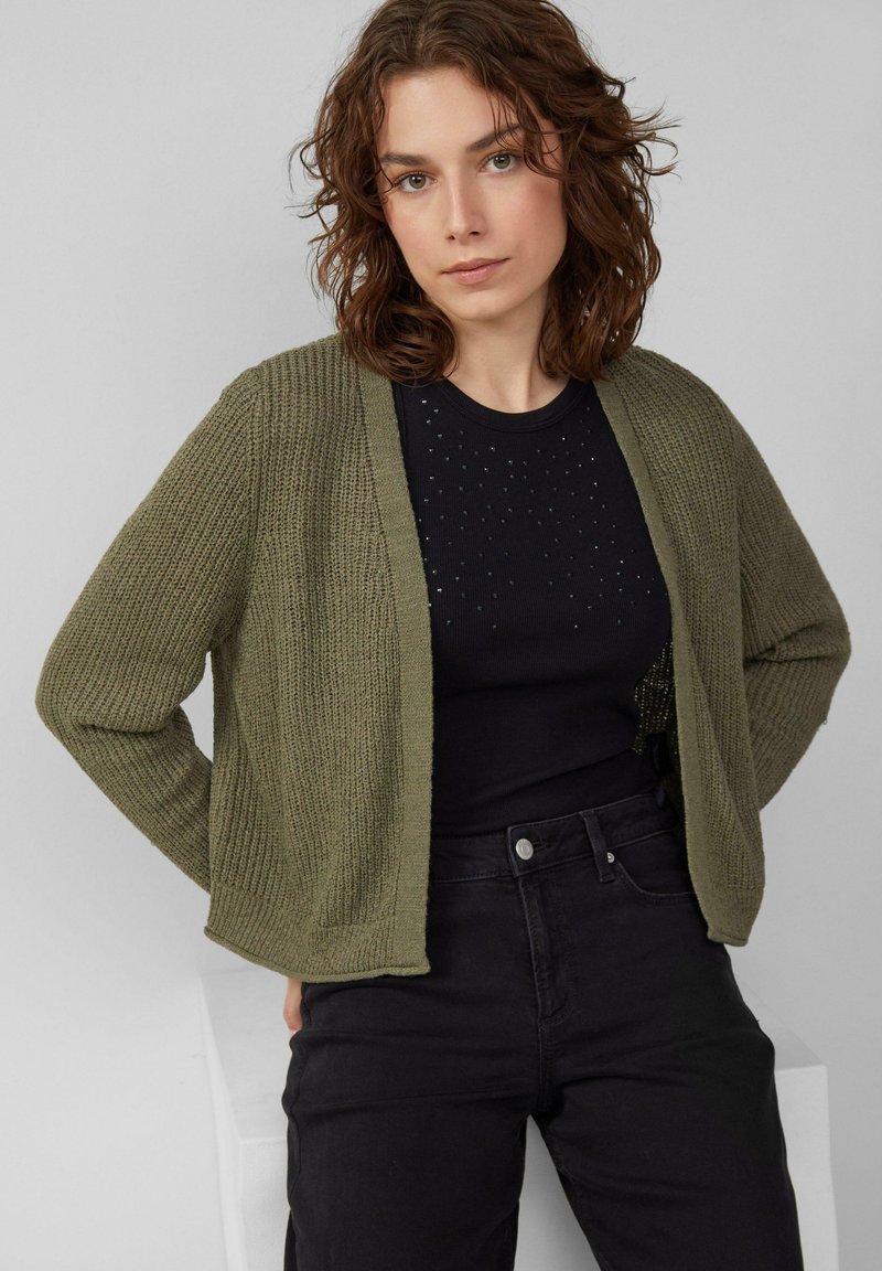 Gilet en tricot vert olive avec une ouverture devant et une texture côtelée, porté sur un haut noir avec de petites embellissements, associé à un jean noir.