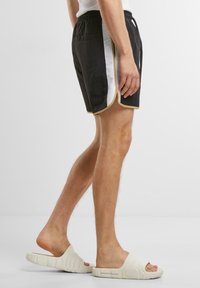 Svarte shorts med hvite og gullstriper på siden, elastisk midje og løs passform. Brukt med lyse sandaler, som viser uformell bekledning.