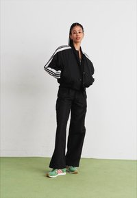 Svart bomberjakke med hvite striper, kombinert med svarte cargo-bukser og grønne sneakers med rosa detaljer. Glatt stoff, avslappet passform.