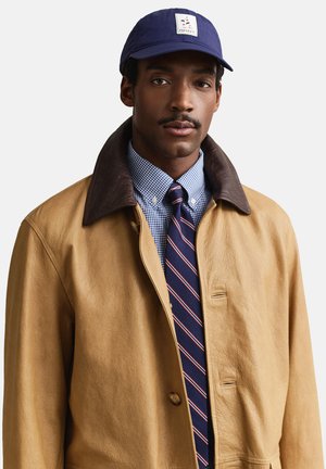 Man draagt een lichtbruine leren jas met een donkerbruine kraag, een blauw geruit overhemd, een gestreepte marineblauwe stropdas en een marineblauwe pet met een zeilbootlogo.