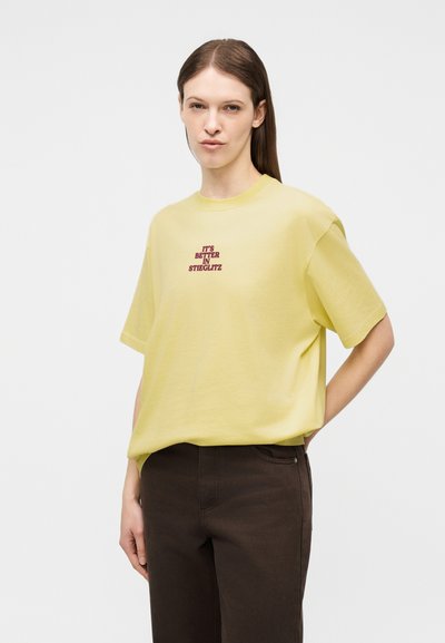 Γυναίκα με ίσια καστανά μαλλιά, φοράει άνετο ανοιχτό κίτρινο μπλουζάκι με κόκκινο κείμενο «IT'S BETTER IN STIEGLITZ» και σκούρο καφέ παντελόνι.