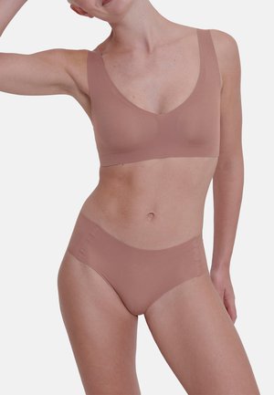 Frau trägt passenden nahtlosen hellbraunen BH und hoch taillierte Slips, posiert mit einem erhobenen Arm vor schlichtem Hintergrund.