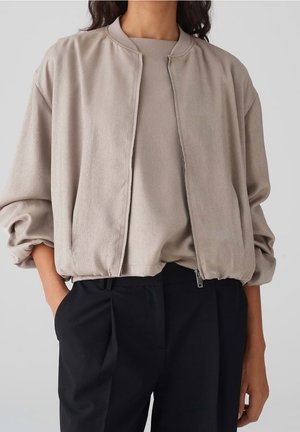 Personne portant une veste bomber beige zippée par-dessus un top assorti avec un pantalon noir plissé, les mains partiellement dans les poches.
