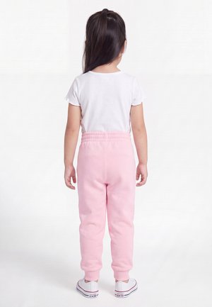 Enfant aux longs cheveux foncés portant un t-shirt blanc à manches courtes, un pantalon de jogging rose clair et des baskets blanches, debout de dos.