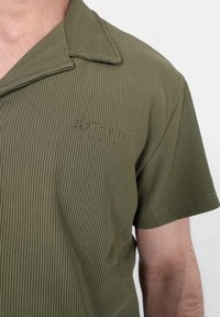Olijfgroene short-sleeve shirt met verticale ribbeltextuur, puntkraag en geborduurd logo op de borst.