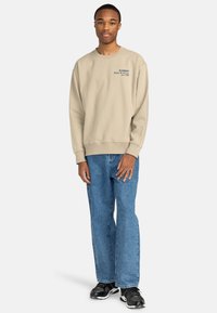 Sudadera beige con cuello redondo y logo bordado en el pecho, combinada con jeans anchos de color azul claro y zapatos deportivos negros.