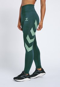 Zielone legginsy z wysokim stanem, z jasnym zielonym wzorem w jodełkę, gładką fakturą i detalem z logo. Noszone z czarnymi butami sportowymi.