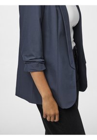 Marineblauer Blazer mit hochgerollten Ärmeln, weicher Textur, offenes Frontdesign und strukturierter Rückseite. Über einem taillierten weißen Oberteil getragen.