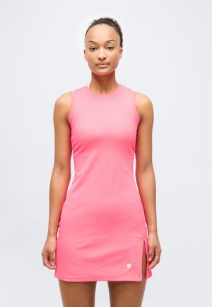 Žena v ružových atletických šatách bez rukávov s bočným rozparkom stojí pred jednoduchým bielym pozadím.
