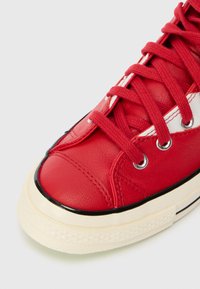 Sapatilha de couro vermelho com biqueira redonda, sola de borracha preta, detalhes em costura branca e atacadores texturizados vermelhos.