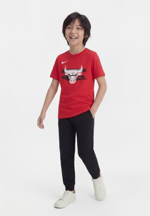 Enfant souriant portant un t-shirt rouge des Chicago Bulls, un pantalon noir et des baskets blanches, debout avec une main dans la poche.