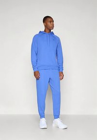 Zils kokvilnas džemperis ar kangura kabatu, saskaņotas sporta bikses un baltas sporta kurpes. Raksturots ar ērtu piegriezumu un vienkāršu dizainu bez logotipiem.