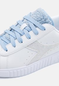 Zapatilla de cuero blanca con tejido azul claro texturizado en el collar, cordones azules y un diseño de estrella plateada brillante en el lateral.