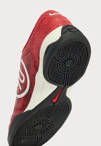 Röd mesh-sneaker med vita accenter, försedd med en texturerad svart gummisula och präglade mönster för grepp. Sidologotyp i vitt.