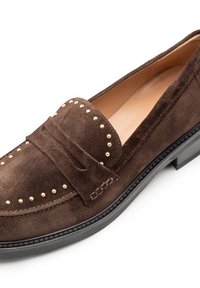 Brauner Wildlederslipper mit goldfarbenen Nieten entlang des Spann, flacher Gummisohle und abgesteppten Akzenten. Glattes Innenfutter ohne sichtbare Markenzeichen.