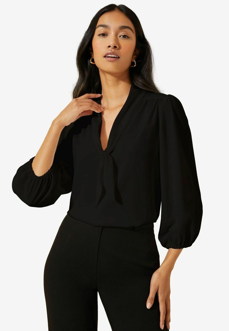 Blusa nera con scollo a V e dettaglio con fiocco, dotata di maniche a sbuffo con polsini elastici, realizzata in tessuto morbido, abbinata a pantaloni neri.