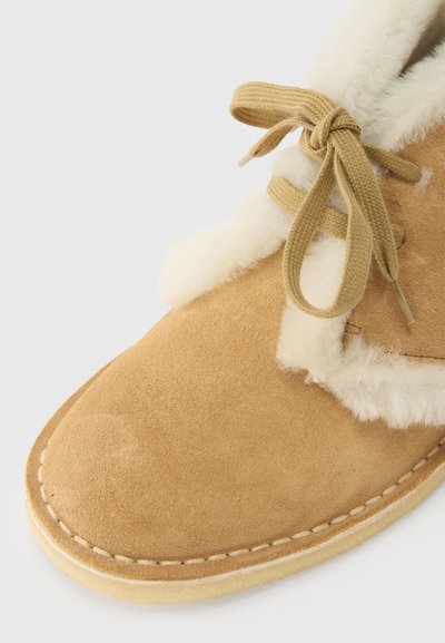 Μποτάκι αστραγάλου από ταμπά σουέτ με λευκή, αφράτη επένδυση και κορδόνια που ταιριάζουν. Διαθέτει στρογγυλεμένη μύτη και εμφανείς ραφές κατά μήκος της σόλας.