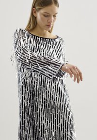 Rochie argintie și neagră cu mâneci lungi, având franjuri metalici verticali și decolteu tip barcă, confecționată dintr-un material strălucitor și ușor.