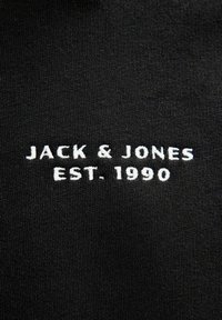 Tela negra con texto bordado en blanco que dice "JACK & JONES EST. 1990." Diseño simple y limpio sin gráficos o patrones adicionales.