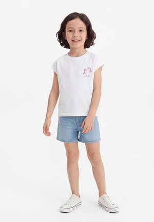 Κορίτσι με κοντά σκοτεινά μαλλιά, φοράει λευκό μπλουζάκι με γραφικό μονόκερο, μπλε τζιν σορτς και λευκά αθλητικά παπούτσια, στέκεται και χαμογελά.