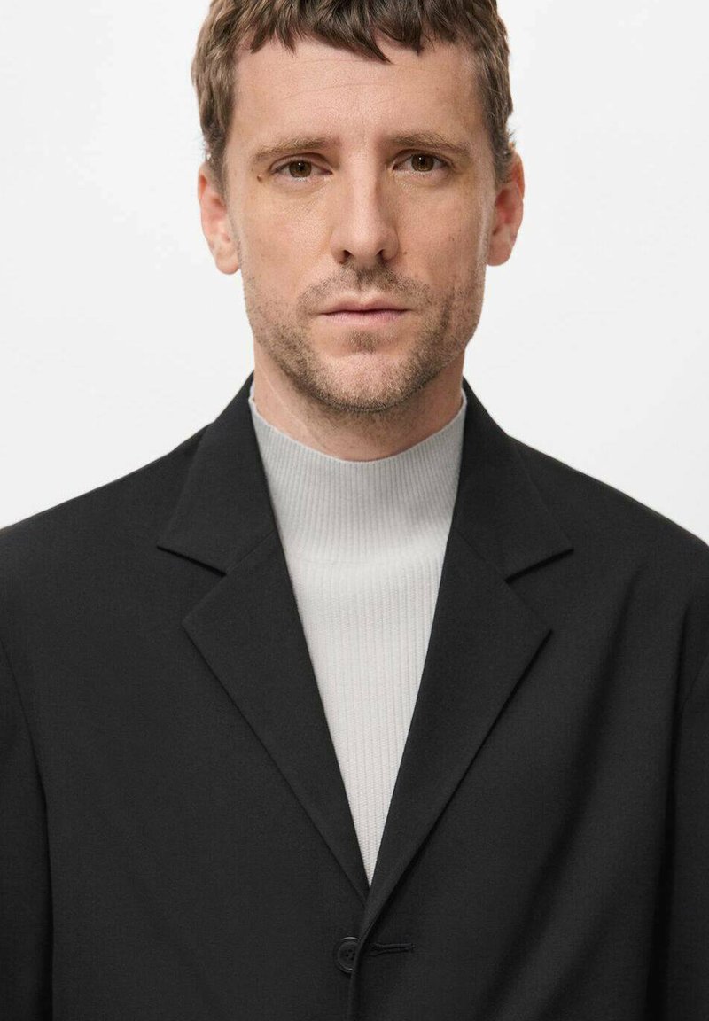 Blazer nero con rever a punta e chiusura a singolo bottone, indossato sopra un dolcevita a coste grigio chiaro, caratterizzato da una texture di tessuto liscia.