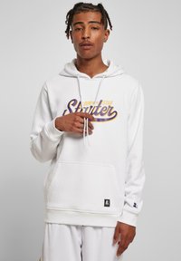 Hoodie blanc avec un texte graphique violet et doré, poche kangourou avant, cordons de serrage, poignets côtelés et un patch logo près de l'ourlet.
