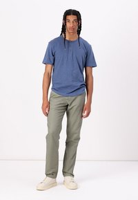 Kortermet blå henley t-skjorte med tre knapper, kombinert med lysegrønne bukser og beige joggesko, stående på en ensfarget bakgrunn.
