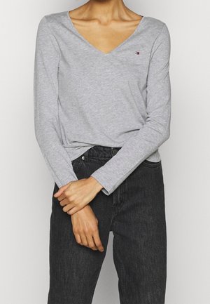 Personne portant un t-shirt gris clair à manches longues et col en V ainsi qu'un jean noir taille haute, les mains croisées à la taille.