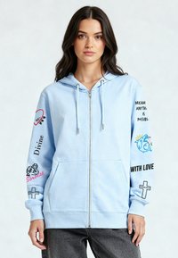 Felpa con cappuccio azzurro chiaro con cerniera e grafiche stampate sulle maniche, incluse parole e simboli. Presenta tasche frontali e cappuccio regolabile con coulisse.