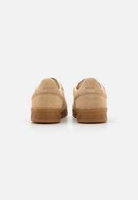 Beige suede sneakers with a textured back, rubber sole, and embossed logo detail. Low-top design and neutral color for versatile styling.