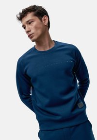 Navy modri pulover s krožnim izrezom, raglan rokavi in embosiranim napisom čez prsa. Gladka tkanina z minimalnimi šivi.