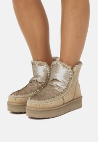 Stivaletti alla caviglia dorati con paillettes, foderati in pelliccia sintetica, base in suede beige e cuciture decorative. Suola piatta con grip testurizzato.